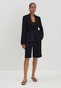 Zwarte, op maat gemaakte blazer met een enkele knoop en zakken, gecombineerd met zwarte shorts tot de knie. Model draagt platte zwarte sandalen.