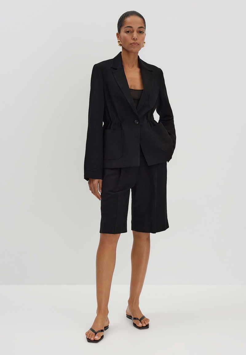 Zwarte, op maat gemaakte blazer met een enkele knoop en zakken, gecombineerd met zwarte shorts tot de knie. Model draagt platte zwarte sandalen.