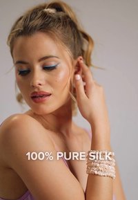 Siden scrunchies i ljusrosa placerade på ett handled, med en mjuk textur. Kvinna i en lilalila topp visar en mjuk, naturlig makeuplook med blå eyeliner.