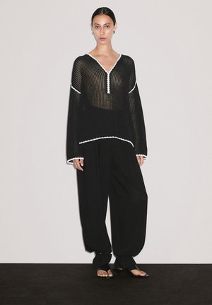 Frau steht vor einem schlichten Hintergrund und trägt schwarze lockere Hosen sowie einen schwarzen Pullover aus offenem Gewebe mit weißen Akzenten, kombiniert mit schwarzen Sandalen.