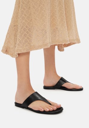 Černé kožené žabky s širokým páskem a plochou podrážkou, spárované s béžovými krajkovými šaty. Povrch je hladký s minimálními detaily.