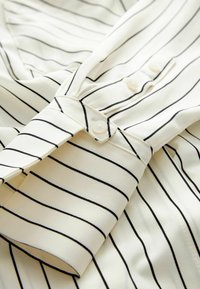 Manche de chemise blanche à fines rayures verticales noires, poignet boutonné et boutons visibles sur la patte de boutonnage.