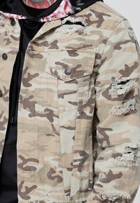 Kamofarbinen denimjakke med beige og brune mønstre, slitte detaljer på ermene, to brystlommer og knappelukking. Teksturert stoff synlig.