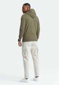 Olivgrüne Kapuzenjacke mit einer großen Kapuze, kombiniert mit hellbeigen Cargo-Hosen mit Seitentaschen und einem Patch, abgerundet mit weißen Sneakern.