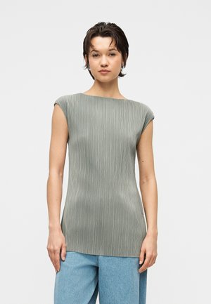 Femme aux cheveux courts et foncés, portant un haut sans manches gris clair côtelé et un jean bleu clair, debout devant un fond blanc uni.