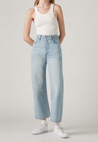 Jeans de tiro alto en un azul claro con un corte de pierna amplia, que presentan un cierre de botón y cremallera, combinados con una camiseta de tirantes blanca de canalé y zapatillas blancas.