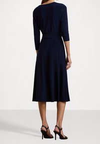 Mujer vestida con un vestido midi azul marino de mangas tres cuartos y un cinturón, combinándolo con sandalias de tacón alto negras, de pie dando la espalda.