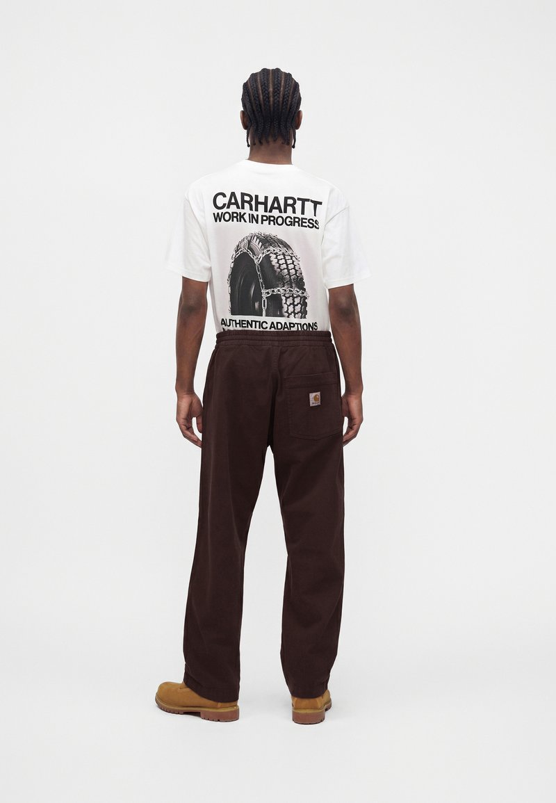 Uomo con capelli intrecciati visto di spalle, indossa una t-shirt grafica bianca Carhartt, pantaloni marrone scuro e stivali da lavoro beige su sfondo bianco semplice.
