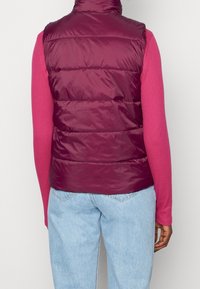 Burgunderfarbene Pufferweste mit hohem Kragen, horizontalen Nähten und glänzender Textur, getragen über einem rosa Langarmtop. Hellblaue Jeans sind sichtbar.