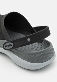 Fekete és szürke slip-on cipő texturált felsőrészsel, gumitalppal és állítható pánttal, amely kör alakú mintázattal rendelkezik a talpán.