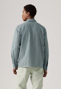 Gestreepte lichtblauwe shirt met een kraag, lange mouwen en knoopaccenten. De stof heeft een gestructureerde uitstraling, gecombineerd met lichtgroene broek.