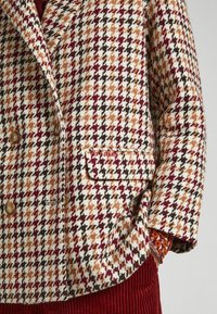 Chaqueta de doble botonadura con un patrón de pata de gallo en burdeos, naranja y negro, con botones marrones y detalle de bolsillo lateral.