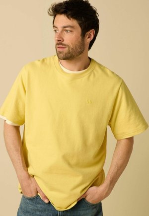 Man met donker haar en baard die een geel sweatshirt met korte mouwen en blauwe spijkerbroek draagt, kijkt opzij met handen in de zakken.