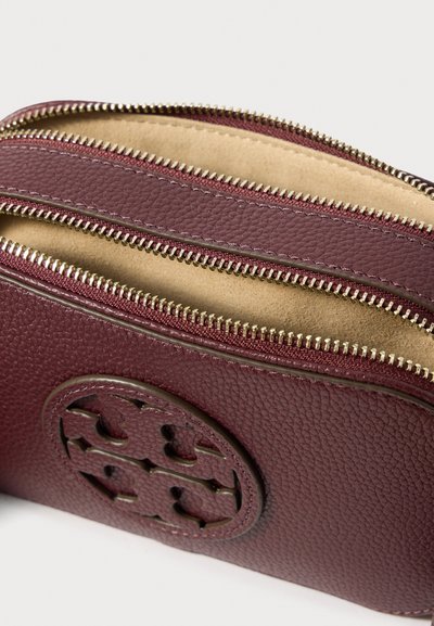 Μικρό μωβ δερμάτινο πορτοφολάκι με δύο ανοιχτές χρυσές φερμουάρ και έναν στρογγυλό διακοσμητικό έμβλημα μπροστά.