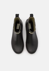 Bottes de cheville en caoutchouc noir avec une finition lisse, dotées d'un intérieur vert, de languettes pour un enfilage facile, et d'une étiquette de marque sur le côté.