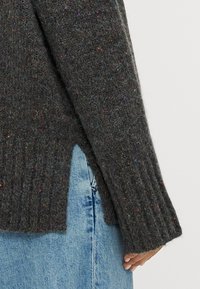 Maglione lavorato a maglia grigio scuro con polsini a costine, caratterizzato da macchie multicolori in tutto il tessuto. Dettaglio con spacco laterale sopra un tessuto denin azzurro chiaro.