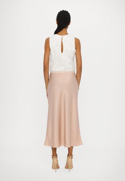 Weiße ärmellose Bluse mit floraler Stickerei und Schlüsselloch-Ausschnitt hinten; kombiniert mit einem blush-farbenen Satin-Midirock und beigen Absätzen.