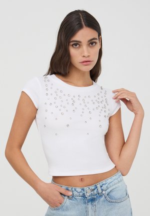 Maglietta cropped bianca a coste con scollo rotondo e maniche a cappuccio. Decorata con borchie argento in un motivo a gradiente sul davanti.