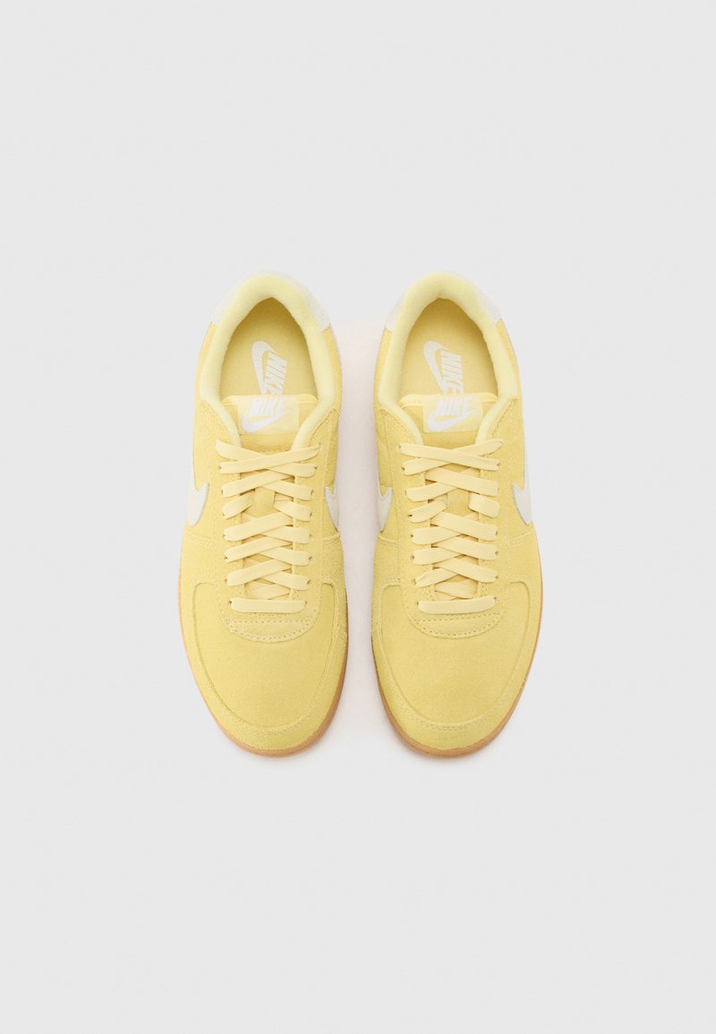 Zapatillas de ante amarillas con acentos en blanco, cordones redondos y una suela de goma texturizada. Presenta un logo en la lengüeta y en el lateral.