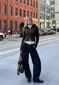 Smeđi rebrasti zip-up pulover s detaljem od umjetnog krzna, bijela košulja ispod i tamne široke traperice. Drži tamnu torbu s krznenim akcentom.