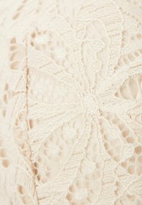 Koronkowy materiał w delikatnym beżowym odcieniu z misternym wzorem kwiatowych motywów i teksturowanymi detalami, pokazujący otwarte i pełne sekcje.