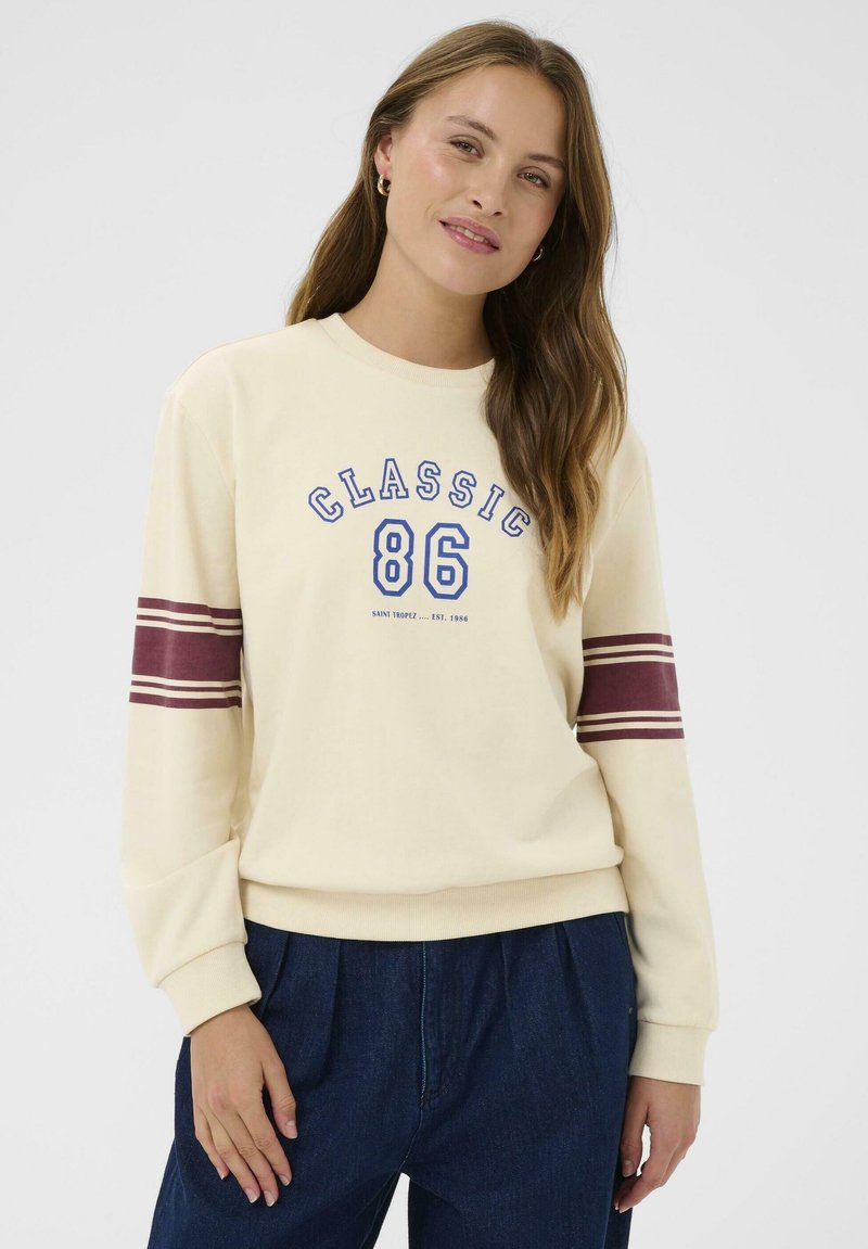 Sudadera crema con texto azul "CLASSIC 86" y rayas color burdeos en las mangas. Tejido suave y corte relajado, combinada con pantalones azul oscuro.