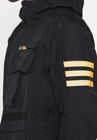 Coupe-vent noir avec une poche frontale, arborant un logo orange et trois bandes horizontales sur la manche. Tissu texturé avec une capuche.