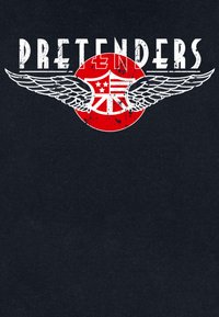 Maglietta nera con la scritta bianca consumata "PRETENDERS" sopra un design circolare rosso con grafiche di ali e un emblema stilizzato.