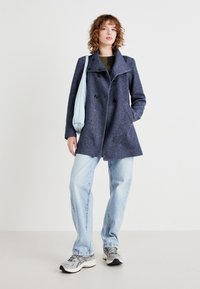Navy blue, double-breasted coat with a high collar, made of textured fabric. Paired with light blue bag, green top, light jeans, and sneakers.