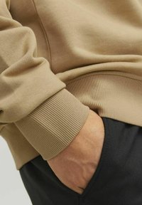 Beige sweatshirt with ribbed cuffs, close-up showing soft texture and a hand in a black pocket, highlighting contrasting colors and materials.