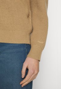 Pull en maille côtelée beige avec une manche retroussée, associé à un jean en denim bleu. Le tissu présente un motif texturé et une coupe légèrement ajustée.