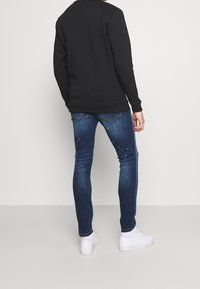Felpa scura con silhouette aderente, abbinata a jeans skinny blu con una finitura sfumata e decorazioni sparse. Sneakers bianche.
