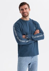 Usmívající se muž s krátkými hnědými vlasy, oblečený v modré košili s dlouhým rukávem a bílým textem na rukávech a světle modrých džínách, s překříženýma rukama.