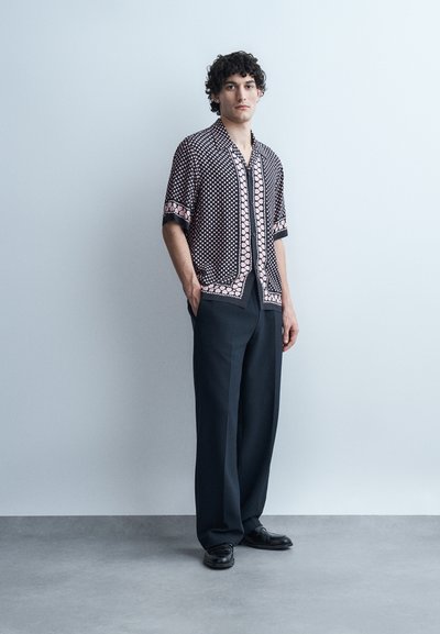 Άνδρας στέκεται με τα χέρια στις τσέπες, φοράει πουκάμισο με μοτίβο και κοντά μανίκια, φαρδιά μαύρη παντελόνια και μαύρα παπούτσια, μπροστά σε γκρι φόντο.
