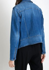 Džínová bunda v středně modré barvě s mírně zkrácenou délkou, s klasickým límcem a zipy na manžetách. Hladká textura a minimální prošití.
