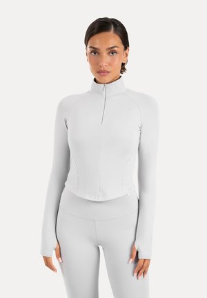 Kvinde iført tætsiddende lysegrå langærmet trøje med lynlås og matchende leggings, stående mod en ensfarvet hvid baggrund.