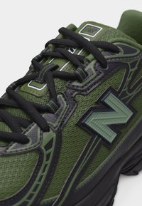 Πράσινο και μαύρο αθλητικό παπούτσι με υφή από δίχτυ, μαύρα κορδόνια και μεγάλο υφασμάτινο λογότυπο "N" στην πλευρά.