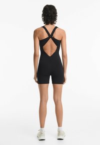 Zwarte geribbelde bodysuit met een V-vormige uitsnijding op de rug en kruislingse straps. Heeft een aansluitend ontwerp en korte benen, gecombineerd met witte sportieve schoenen.
