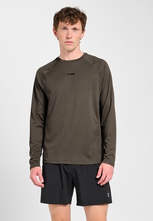 Młody mężczyzna zwrócony przodem, ubrany w ciemnobrązową koszulę z długim rękawem i czarne sportowe szorty na jednolitym jasnym tle.