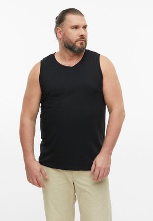 Man met baard en naar achter gebonden haar, gekleed in een zwart mouwloos shirt en beige broek, staand en zijwaarts kijkend.