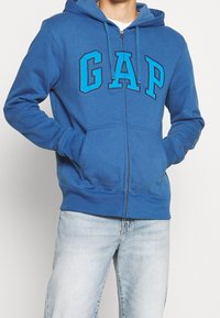 Homme portant un sweat à capuche zippé bleu avec "GAP" en grandes lettres sur la poitrine et un jean délavé clair, mains dans les poches.