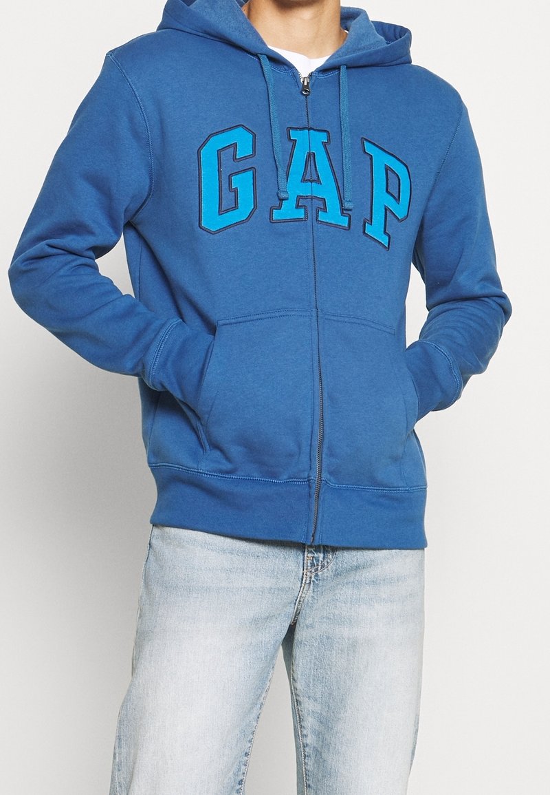 Homme portant un sweat à capuche zippé bleu avec "GAP" en grandes lettres sur la poitrine et un jean délavé clair, mains dans les poches.