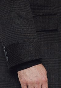 Wełniany płaszcz w ciemnym brązie z delikatnym wzorem w kratkę. Posiada mankiet z trzema czarnymi guzikami i prążkowaną czarną podszewkę.