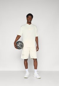 Cremfarvet atletik sæt bestående af en kortærmet trøje og shorts, tekstureret stof og et hævet logo. Model der holder en basketball.