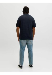 Homme aux cheveux tressés, debout de dos, portant une chemise à manches courtes texturée de couleur foncée, un jean bleu clair et des baskets noires.