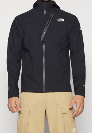 Homme portant une veste North Face noire zippée avec une poche poitrine et un short cargo beige, debout devant un fond uni.