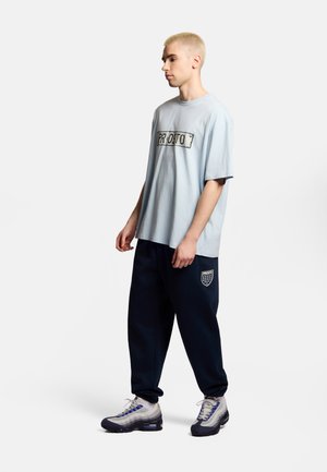 Młoda osoba z krótkimi blond włosami, ubrana w jasnoniebieską ponadgabarytową koszulkę i granatowe spodnie dresowe oraz sneakersy, stojąca na jednolitym tle.