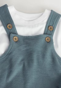 Tricou alb cu guler rotund și nervuri, sub salopetă albastră texturată cu patru nasturi de lemn pe bretele și panoul pieptului.