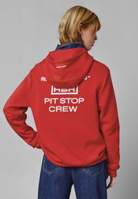 Rdeča kapuca s belim napisom "PIT STOP CREW" na zadnji strani, zaključena s sprednjim žepom in rebrastimi manšeti, nosi se čez modre kavbojke.