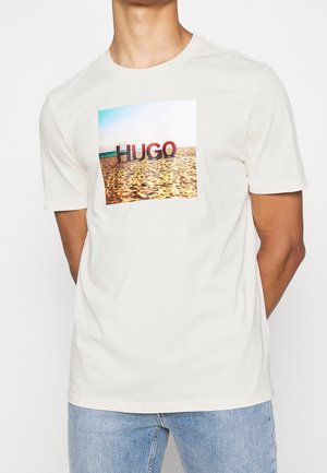 Mann trägt ein weißes T-Shirt mit einer rechteckigen Strandszene und dem Text "HUGO" auf der Brust, kombiniert mit hellblauen Jeans.